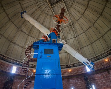 A very large, thin barrelled telescope sits at a slanted angle atop a blue trapezoidal mount. The mount is nearly three stories tall and has a spiral staircase on the left hand side. The photo is taken inside of a giant dome, which can be seen covering the entire background.