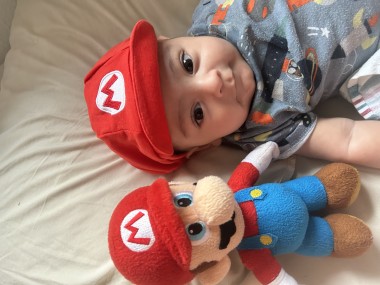 A cute baby in a Mario beanie next to a Mario plush