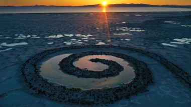 An image of the Earthwork "Spiral Jetty" by Robert Smithson, with the setting sun in the background reflecting off of a pool of water, collected within the spiral.