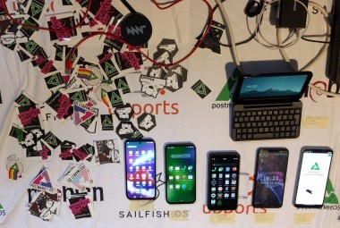A lot of postmarketOS stickers, and phones running postmarketOS: a PinePhone with a keyboard + Plasma Mobile 5, two Fairphone 5 running Plasma Mobile 6 and Gnome Shell on Mobile, a Librem 5 running Phosh, a PinePhone running Sxmo and a Fairphone 4 running Phosh