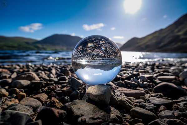 Lensball glass globe photographed on lake shore surrounded by pebbles with lake and mountains in background and reflected inversely in ball