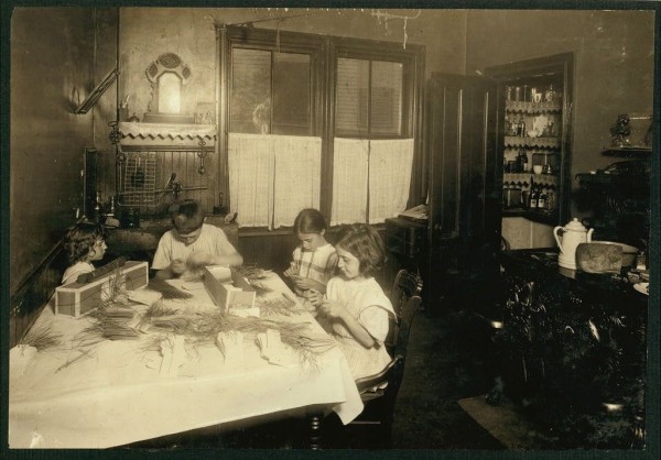  The image shows a black and white photograph of an indoor scene that appears to be from the early 20th century. It depicts three individuals, likely a family, engaged in some form of craft or work at a table covered with cloth. The person on the left is seated, facing the others, while the two individuals on the right are standing and facing away from the camera. They seem to be working together on a task involving fabric, possibly making tags for garments, as indicated by the text description.

The setting suggests a domestic environment with traditional furnishings such as chairs and a dresser visible in the background. The style of clothing and interior decoration is consistent with mid-20th century America. It's important to note that this description is based on the information available and does not include any assumptions about the individuals' identities or personal stories beyond what can be discerned from the image itself.