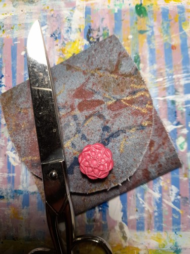 Folded pocket shrine with scissors as weight. The shrine has a textile outside in grey-blue with brown, blue and golden stains looking like abstract watercolours. A pink button with a geometrical flower texture for closure.