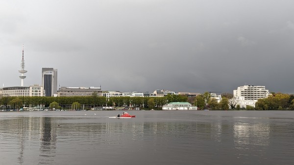 Foto auf die Binnenalster Hamburg. Im Hintergrund rechts: Der markante Fernsehturm. Auf dem Wasser ist ein einzelnes rotes Tretboot zu erkennen. Grauer Himmel über Hamburg.
