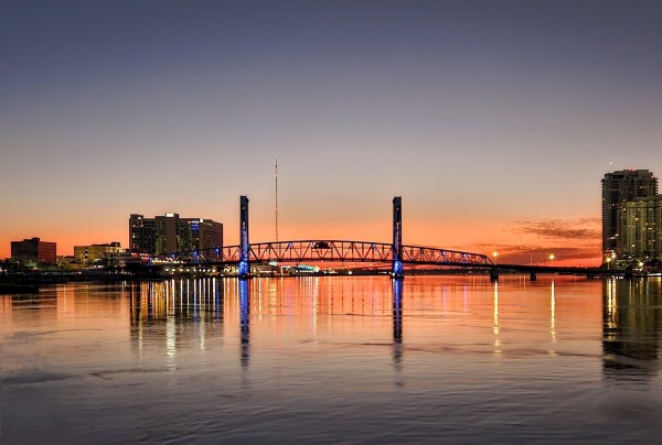 A bright red and orange glow rises at the horizon prior to the sun's arrival for the morning sunrise.  The  bright colors illuminate the city skyline and their colorful lights including a large blue drawbridge, all reflecting their colors and image upon the calm river below.