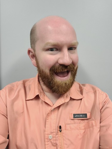 Selfie of a balding, aggressively white man with a beard wearing a pink, short sleeved button up shirt against a light colored wall. He's smiling broadly with his mouth open.