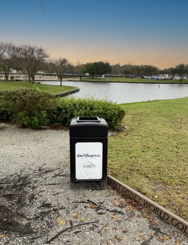 A trash can with the Walt Disney World logo in a park-like setting with a body of water, pathways, and a soft sunset sky in the background. 