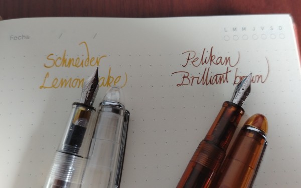Yellow and brown ink writing samples on dot grid paper. Two clear uncapped fountain pens, one colorless and one brown-orange