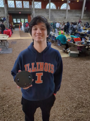 A sleep-deprived young man in an Illinois hoodie holding a black circular 1 pound full body spinner robot