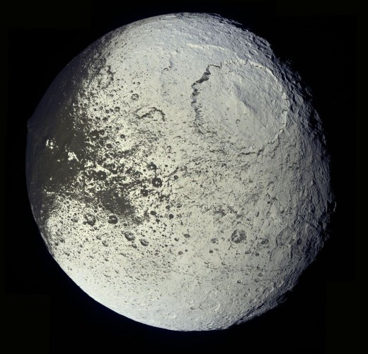 The brightly lit sphere of Iapetus as seen by NASA's Cassini spacecraft. The surface is mostly white, with an area of dark grey to the upper left and two large craters overlapping on the upper right.
