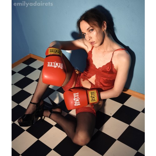 Emily Adaire sitting on a black/white checkered floor, leaning against a blue wall. She is wearing red satin lingerie, stockings and black high heels, as well as red boxing gloves. She looks tired into the camera.