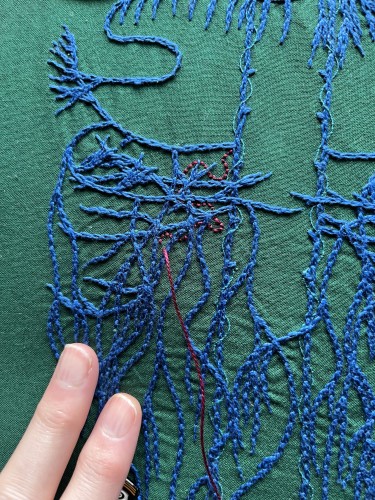 A close up of an embroidery focused on the abdominal vagus stitched in blue chain stitch in perlé thread on dark green cotton. There are many layers of crossing wavy lines. Spiralling around the very straight main vagus is single strand whipped back stitch in electric blue. Some roiling burgundy back stitch in burgundy perlé thread has been started in the left side of the abdomen. 