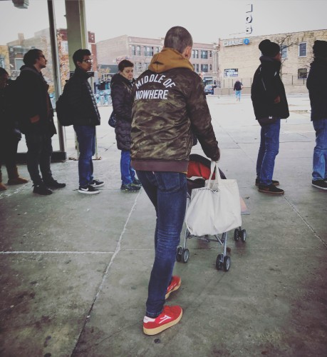 A man wearing a camouflage jacket with the words “MIDDLE OF NOWHERE” written on the back in white all caps letters. He is pushing a fold-up stroller through a queue of people waiting for the bus. 