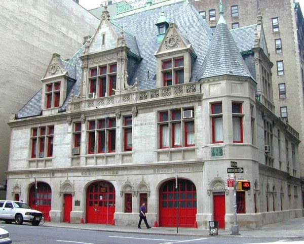 A multi-story French style building with white walls, gray tall sloped roof, red window frames, and large red doors on the first floor.