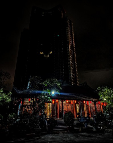 A late night photo of a glowing temple with a highrise under construction behind it. The temple is all lantern-lit, dark wood with gold-leaf sino-vietic characters and interior panels shining through open doors and windows. It is a single floor with wide stairs leading up to the entrance. A massive incense urn sits at the foot of the stairs. The highrise apartment building behind rises 30 floors and is mostly dark. What little is lit is dominated by a single, vertical band of lights.
