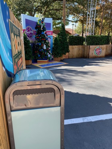 Outdoor scene featuring a Christmas tree with decorations, surrounded by smaller trees and signage, adjacent to a parking area with a trash can in the foreground. 