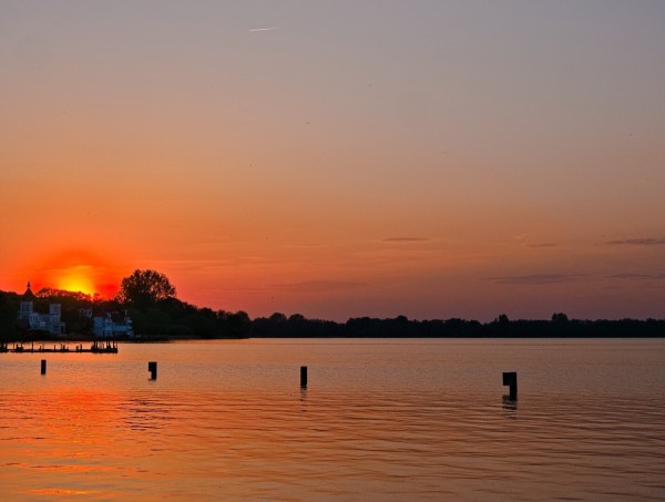 A large lake in the foreground. The water is orange, just like the sun, which is setting behind a white house on the other shore. The sky is drenched in orange and purple.