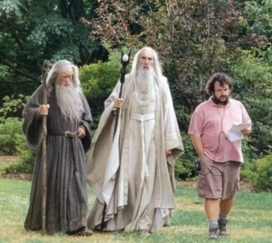 Two actors as wizards in Lord of the Rings - in full costume with beards and staffs - listening while walking to director Peter Jackson casually wearing a polo shirt and shorts.