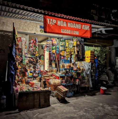 A late evening photo of a neighborhood tạp hóa (mom and pop convenience store) burning bright on an otherwise darkened street in Hanoi. A red banner with white text hangs from a corrugated metal extension over the front stoop. it reads, "TẠP HÓA HOÀNG ANH"  and lists a phone number. Goods spill out onto the sidewalk and street from the first floor shop. We see cases of beer stacked on the right next to bags of snack chips and dried fruit. Candies and cakes are on the left. Single serving packets of Milo hang from a rack with other offerings. Boxes of instant noodles form a makeshift wall along the entry aisle to the right, and a glass-topped ice cream freezer is tucked away behind to outward facing racks. Above the door, just around the edge of the banner, we can make out and blue and white address indicator that reads, "17B."