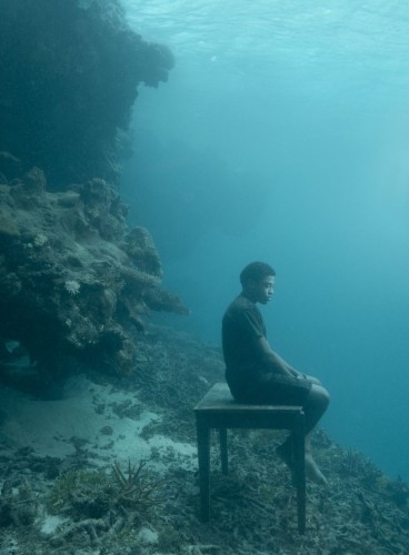 Photography. A color photo of a man of color sitting on a wooden table underwater in the sea. The photo is blurred blue/ turquoise and semi-dark. The young man can be seen from the side. He is wearing shorts and a T-shirt and is sitting there with his eyes open, surrounded by an underwater world of seaweed, sand and coastal cliffs. 
Info: The photo is part of a series (The Day May Break) that focuses on the inhabitants of the South Pacific islands affected by rising oceans due to climate change.
The locals in these photos, taken in the ocean off the coast of Fiji, are representative of the many people whose homes, land and livelihoods will be lost in the coming decades due to rising waters.