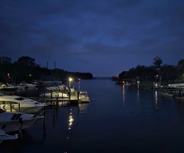 Beneath a dark stormy sky a waterway appears calm, reflecting a variety of pier and boat lights upon the glass like surface.  Many shades of blue between the dark sky and calm waters.