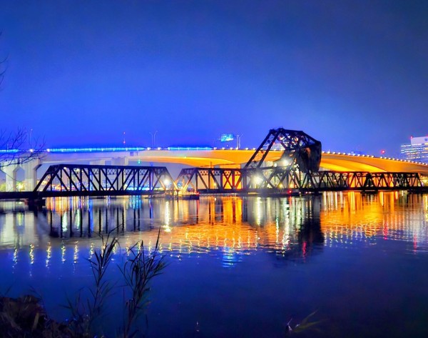 Between the night and approaching sunrise, a blue glow overtakes the sky above a calm, equally blue river where a downtown skyline is illuminated with numerous bright colorful lights as a backdrop to bridges crossing the river. In the background a large concrete bridge with bright blue and yellow lighting shines on the adjacent railroad drawbridge trestle, rusty and black, now in the down position as a train approaches, all casting reflections on the calm waters below.