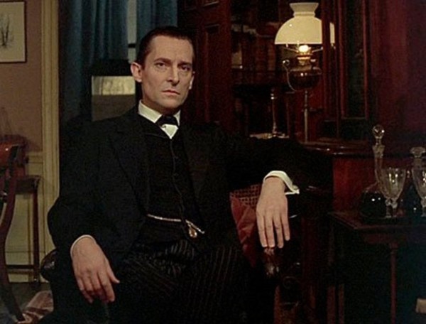 Jeremy Brett as Sherlock Holmes. He’s wearing a dark suit with starched collar and tie, and a vest. A watch chain can be seen leading to his vest pocket. He’s relaxing on a chair in a Victorian era sitting room, a crystal decanter set beside him and an oil lamp behind him. 