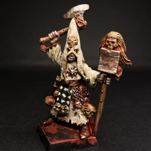 Cult of the Possessed Miniature from Mordheim. Painted in off white creams, reds and blacks. This one is holding an axe above its head and holding a staff with a skull on it, a piece of paper is nailed to the staff. 