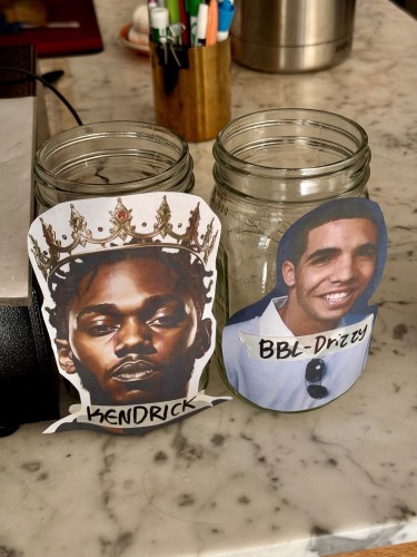 Two tip jars on a coffee shop counter. One has a photo of Kendrick on it, the other has a photo of Drake. It is not clear in this photo which one has more money in it. 