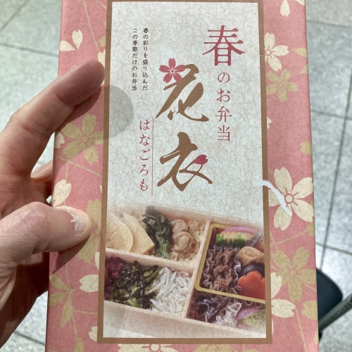 Hand holding a booklet with Japanese text and an image of a traditional Japanese bento box. The booklet has a pink floral design.