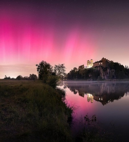 Photography. A wonderful color photo of the glow of the aurora borealis over the Benedictine Abbey in Tyniec, Krakow on 10.05.2024. In the night shot you can see the grass-covered banks of the Vistula (river) and the old abbey on an island. Above the river, the sky glows with pink and light yellow rays. The lights are reflected in the water. A wonderful nature experience