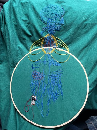 An image of an old engraving of a vagus nerve is embroidered on green cotton. The gut is in a large bamboo embroidery hoop. The embroidery of the main nerve lines is done in blue chain stitch in perlé cotton. The brain and some of the face is stitched in the same thread in backstitch. Details in the face are stitched in single strand floss. A bright red anatomical heart is stitched in two and one strand red whipped back stitch. Around it a bright golden yellow halo in a vesica piscis shape is stitched in single strand thread. Three sets of ribs have are stitched in bright green/yellow stem stitch. There are some tiny details in different colours along some of the nerves. Some burgundy roiling is being stitched in the left gut area. A needle minder in the shape of an hourglass that reads, "This is taking forever," sits near the stitches holding a needle.