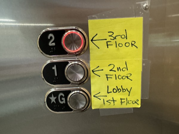 Elevator buttons. Yellow post its are taped next to the buttons. Button 2 is labeled 3rd floor; 1 is labeled 2nd floor, and G is labeled Lobby 1st Floor. 