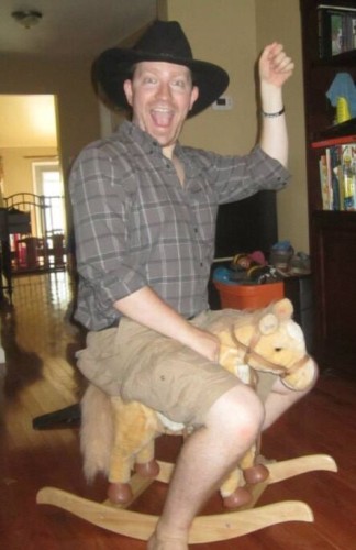 Photograph of a bearded, white man wearing a black cowboy hat, black checkered shirt, and khaki shorts riding on a small children's wooden rocking horse with their left hand elevated in the air.