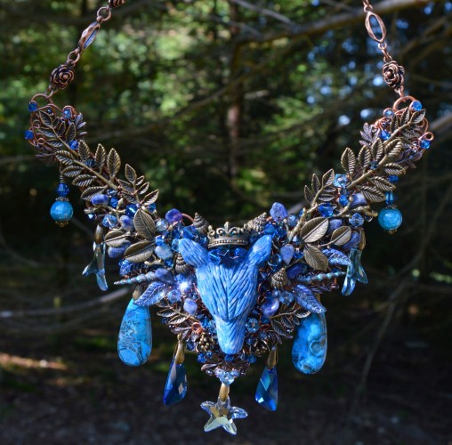 A large elaborate V-shaped necklace wrought in wire, and densely woven with tiny sapphire blue crystals and beads. In the centre is a wolf had carved from rich blueLapis Lazuli Stone. He wears a bronze crown, and a rich patten of bronze leaves and blue beads branches either side of him. Below hang a starfish and clear blue Swarovski crystal charms in various shapes. He hangs from a chain worked with roses, aghainst a dark forest background.