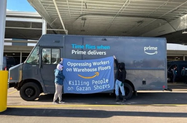 Two activists hold a banner against an Amazon delivery van.  Blue background with a yellow "Amazon smile" in the middle, text reads
OPPRESSING WORKERS ON WAREHOUSE FLOORS
KILLING PEOPLE ON GAZAN SHORES