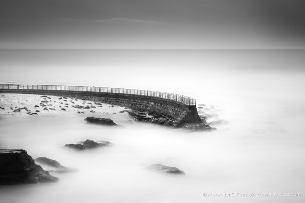 A black & white long exposure photo showing a seawall curving into the frame from the left. Some coastal rocks also stick out of the water. The surf is rendered completely smooth and silky by the slow shutter speed.