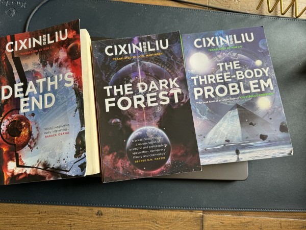 Three books from Cixin Liu's "Remembrance of Earth’s Past" trilogy are displayed on a table, with artistic cosmic-themed covers, from left to right: "Death's End," "The Dark Forest," and "The Three-Body Problem
