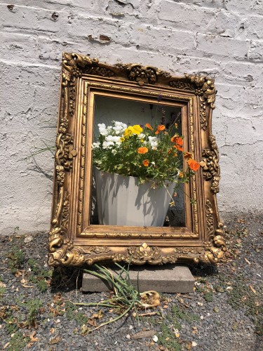 À planter full of orange and yellow flowers sits against a wall. A mn elaborate gold picture frame has been leaned against the wall, surrounding the planter. 