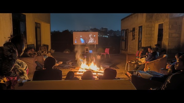 Sofas are arranged in a semi-circle around a bonfire, facing a projector screen (in the background of the image) on which is a scene from Star Wars, A New Hope. The scene is set in a compound and buildings are visible on the sides of the image. The night sky is a dark silver colour (but might be another colour, I'm not good at describing those) 
