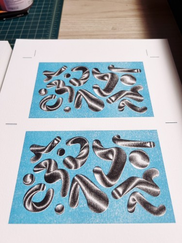 Squiggly metallic looking shapes Risograph printed in aqua and black colors on a sheet of white paper on a light-colored wooden surface