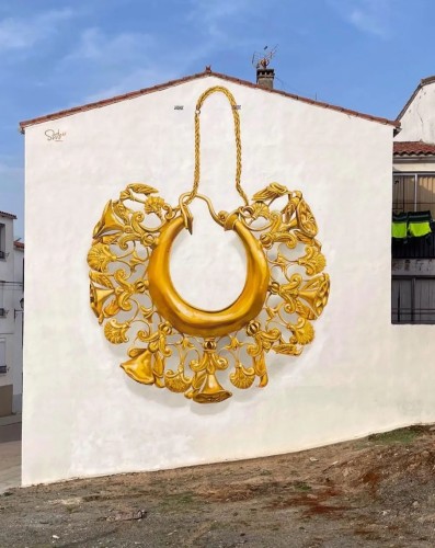 Streetartwall. On the rear outside wall of a one-story house on a hillside, a mural of a richly decorated, old, golden earring has been sprayed/painted in a realistic painting style. The golden earring consists of a thin gold chain as a hanger and an inner golden ring around which small bells, ornaments and leaves are arranged. The wall painting is very realistic and has a 3D effect with shadows.
Info: The treasure trove of Aliseda (Spanish: Tesoro de Aliseda) is a treasure consisting of several large and over 300 small, finely crafted pieces of gold jewelry. It is dated to the beginning of the Iron Age (700 to 250 BC). Only copies can be seen in the city. The treasure itself is located in the "Museo Arqueológico Nacional de España".