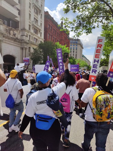 This is a picture of a multi-racial and multi-gendered group of people marching in Washington DC in support of clemency for incarcerated women. They are carrying signs that say FreeHer, as well as signs with the names of individual women who are incarcerated and some State banners as well. It's sunny outside and the marches are in the street in DC walking down towards Freedom Plaza.