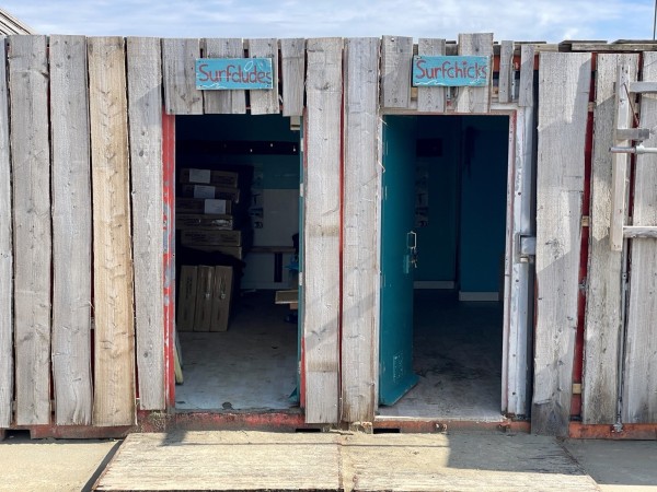 Wooden beach huts with open doors, one labeled "Surfdudes" and the other "Surfchicks," revealing storage space inside.