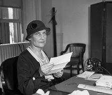 Posed photo of Nellie Tayloe Ross at her desk at the Mint. She is a white woman in a dark cloche hat.