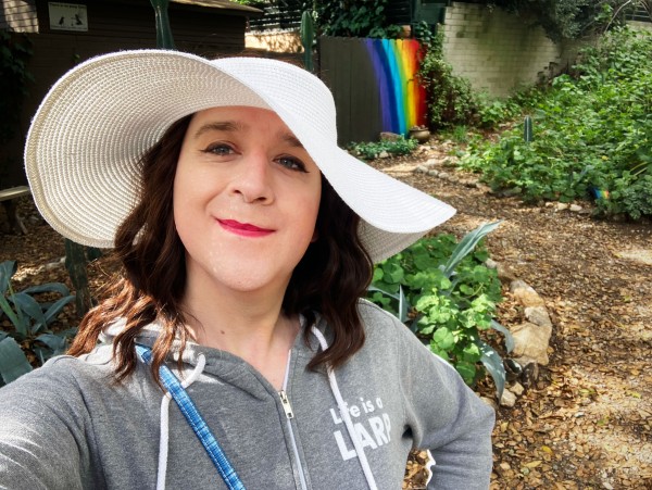 A woman with pale skin and brown hair is wearing a hoodie and big floppy white hat outdoors, smiling at the camera. She is in a glen of trees, standing in front of a brown fence with a rainbow painted on it
