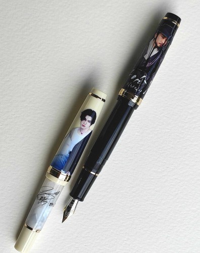 Two fountain pens with images printed on them. The black fountain pen on the upper right features a Kdrama actor in period costume. The smaller cream fountain pen on the lower left features a Kpop idol.
