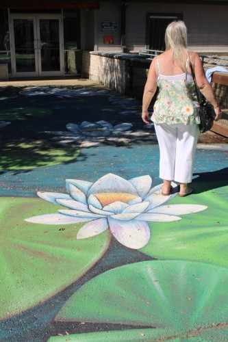 Woman with light blond hair in a summer top with large daisies and white pants with a black purse over her right shoulder facing away from the camera standing on a mural of o lotus and lily pads on a walkway in front of a building.