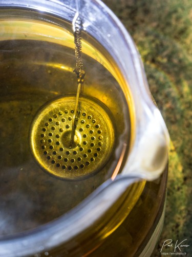 Photo en format vertical, en contre-plongée sur un récipient rempli d'eau colorée par le contenu d'une boule à thé suspendu à une chaîne accrochée au rebord du récipient.