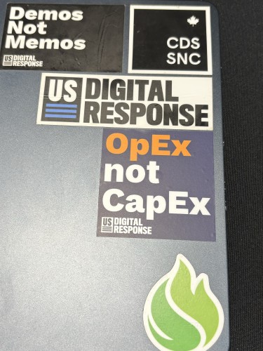 A close-up on a MacBook case, showing several stickers. Centered is one that says “OpEx not CapEx.” Also seen:

“Demos Not Memos”
“US DIGITAL RESPONSE”
“CDS / SNC”
